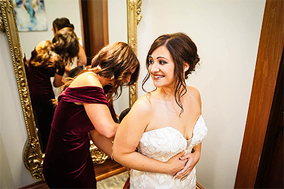 Bride smiling with another woman helping her fasten the back of her wedding dress in front of full length mirror